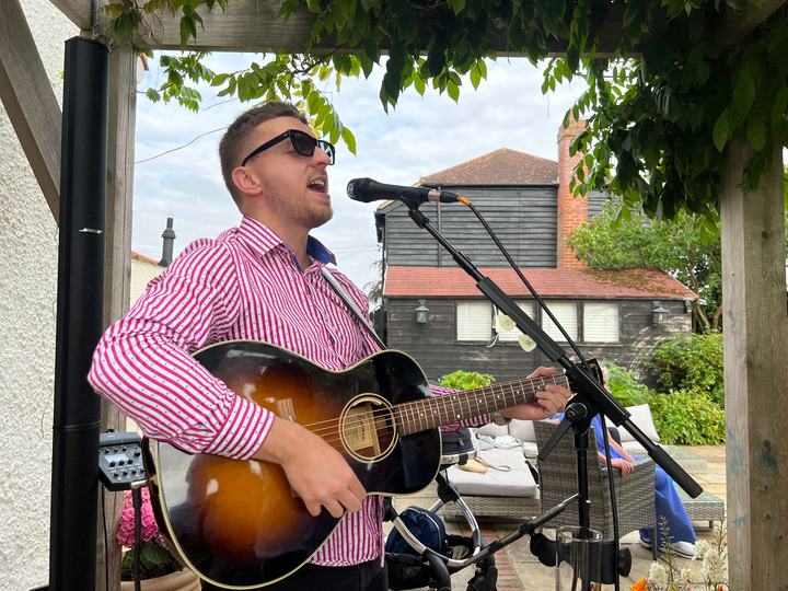 Tom Ryder playing acoustic guitar outdoors under a pergola at a wedding